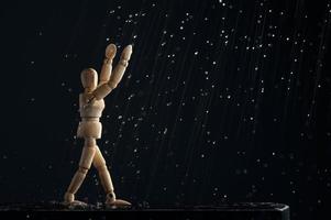maniquí de madera de un hombre bajo la lluvia levanta las manos sobre un fondo negro. el concepto de alegría. una figura de madera camina a través de charcos. foto