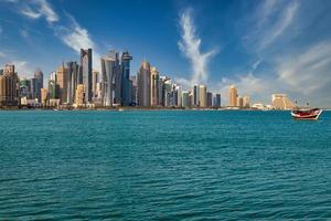 Vista de la luz del día del horizonte de Doha Qatar con nubes en el cielo que muestran rascacielos y dhow con la bandera de Qatar en el fondo y el Golfo Arábigo en primer plano foto