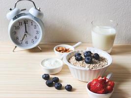 breakfast set, oat or granola in white bowl, fresh blueberries and strawberries, a  glass of milk and white vintage alarm clock 7 am.  on wooden table. Healthy breakfast concept. photo