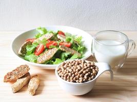 semillas de soja en tazón blanco, tempeh en plato de ensalada y leche de soja en mesa de madera, productos de soja. foto