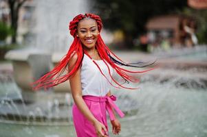 chica afroamericana de moda en pantalones rosas y rastas rojas posadas al aire libre contra fuentes. foto