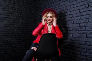 retrato de estudio de una chica rubia con sombrero rojo, gafas y chaqueta de cuero posada en una silla contra una pared de ladrillo. foto