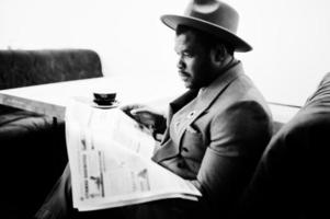 elegante modelo afroamericano con chaqueta gris, corbata y sombrero rojo, bebe café en la cafetería y lee periódicos. foto en blanco y negro.