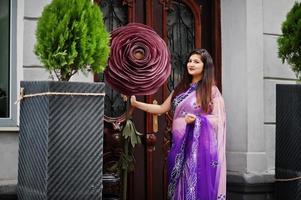Indian hindu girl at traditional violet saree posed at street near big decorated rose. photo