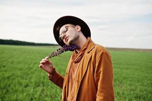 Stylish man in glasses, brown jacket and hat posed on green field. photo