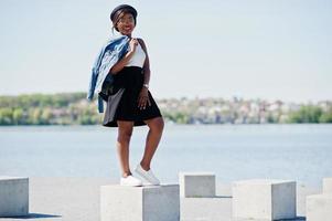 Stylish african american model in glasses hat, jeans jacket and black skirt posed outdoor. photo