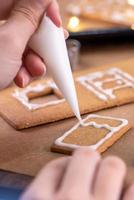 Young woman is decorating Christmas Gingerbread House cookies biscuit at home with frosting topping in icing bag, close up, lifestyle. photo