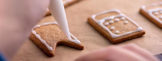 mujer joven está decorando galletas de casa de pan de jengibre de navidad en casa con cobertura de glaseado en bolsa de hielo, primer plano, estilo de vida. foto