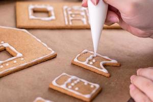 Young woman is decorating Christmas Gingerbread House cookies biscuit at home with frosting topping in icing bag, close up, lifestyle. photo