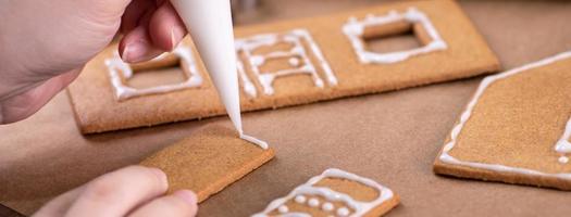 Young woman is decorating Christmas Gingerbread House cookies biscuit at home with frosting topping in icing bag, close up, lifestyle. photo