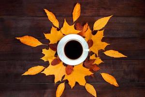 Cup of hot strong coffee and fallen leaves on wooden background. Hello autumn concept. Warm beverage for cold weather. photo