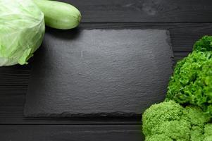 Black stone board on a black wooden background decorated with green vegetables and herbs. photo