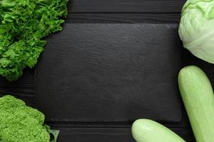 Black stone board on a black wooden background decorated with green vegetables and herbs. photo