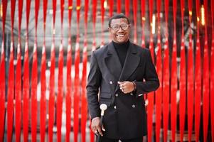 Stylish african american gentleman in elegant black jacket, holding retro walking stick as cane flask or tippling cane with golden diamond ball handle. Rich fashionable afro man against red wall. photo