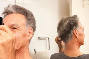 hombre maduro 60plus peinando su cabello mayormente gris en la parte posterior de su cabeza frente a un espejo foto