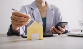 The hand is pressing calculators, piggy bank with wooden house. buy or rent question on note with calculators on desk. Save money and buy house concept. photo