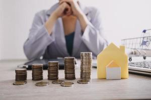 The hand is pressing calculators, piggy bank with wooden house. buy or rent question on note with calculators on desk. Save money and buy house concept. photo