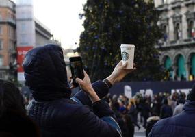 Milan, Italy - December 14, 2019 - A young man holds up a Starbucks paper cup and takes a photo of it with his mobile phone.