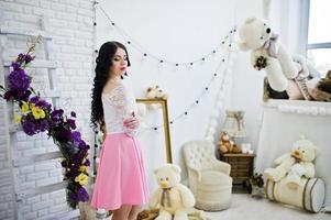 jovencita morena con falda rosa y blusa blanca posada en el interior contra la habitación con oso de juguete. foto