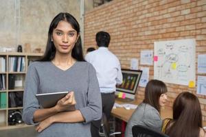 retrato de una joven mujer de negocios asiática que sostiene una tableta digital en un lugar de trabajo de oficina creativo. equipo en reunión en segundo plano foto