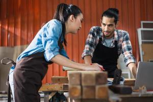 Young Asian couple carpenters man and woman discussing about design of products on laptop in woodcraft carpentry workshop. photo