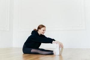 Fit redhead woman stretches muscles on legs, has sporty body, focused into distance, looks happily, wears black clothes, poses on floor in spacious hall. People, flexibility and sport concept photo