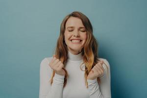 Excited young lady clenched fists waiting for miracle with eyes closed, isolated on blue background photo