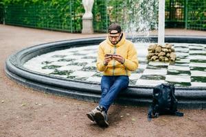 Young attractive bearded man in casual clothes sitting near stone fountain being concentrated into screen of his smartphone watching films while having rest in cozy green park. Man chatting outdoors photo