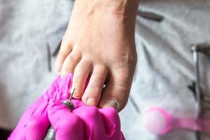 el maestro pedicurista con guantes rosas corta la cutícula y las uñas de los pies de goma laca en el salón de pedicura con un taladro. pedicura profesional en clínica de cosmetología. higiene de los pies en el salón de belleza. foto