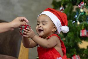 African American baby is happily smiling while receiving little gift box from parents while dressing in christmas dress and santa hat with christmas tree on the back for season celebration concept photo