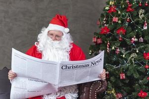 Santa Claus is reading Christmas news from the newspaper while sitting on the sofa couch by the christmas tree for long holiday economics situation and accident traffic transport report information photo