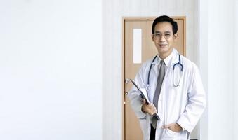 Portrait of asian male doctor at the hospital ward in front of patient room holding clipboard with medical record for better nursing care plan concept photo