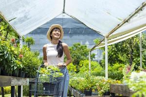 un joven cliente asiático está eligiendo una planta exótica del vivero del centro de jardinería local con un carrito de compras lleno de plantas de verano para la jardinería de fin de semana y el concepto al aire libre