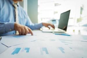 Handsome young businessman working with laptop while sitting on the desk in office and financial statistic graph, blurred background photo