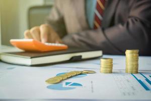 businessman using calculator with stacked coins arranged at office desk and many document data graph in morning light, business concept. photo