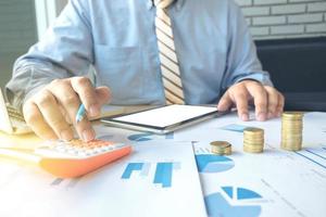 businessman using calculator with stacked coins arranged at office desk and many document data graph in morning light, business concept. photo