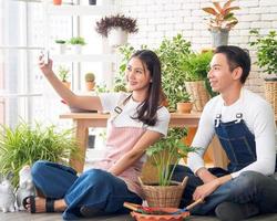 Couple gardener young Asian man woman two person sitting with floor smiling looking hand holding Smartphone selfie camera in calm work shop home plant white wall. hobby job happy and care concept photo