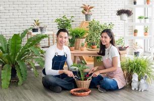 jardinero joven hombre asiático mujer dos personas sentadas con suelo sonriendo mirando mano sosteniendo cesta olla pequeño árbol hoja verde en calma taller casa planta pared blanca. hobby trabajo feliz y cuidado concepto foto