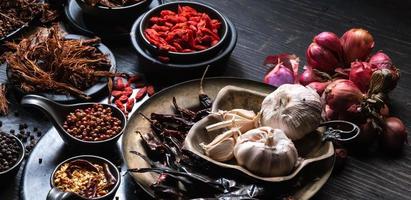 foto de vista superior Se colocaron montones de ajo, cebollas, pimientos, chile y hierbas secas en platos y tazas sobre un fondo de madera negra. se dividió para preparar para cocinar comida india comida saludable