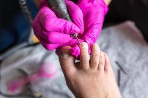 Pedicurist master in pink gloves cuts the cuticle and shellac toe nails in the pedicure salon using drill.  Professional pedicure in cosmetology clinic. Hygiene for feet in beauty salon. photo