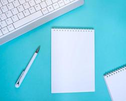 An empty notepad with a pen is on a blue background with a laptop and supplies. Top view with copy space, flat lay. photo
