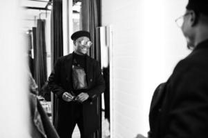 Stylish casual african american man at black beret and overcoat with waist bag at fitting room clothes store, looking on mirror. photo