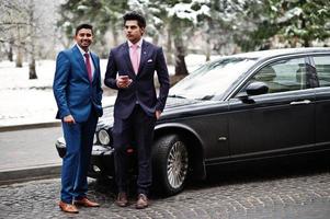 dos elegantes modelos indios de moda en traje posaron en el día de invierno contra un auto clásico negro. foto