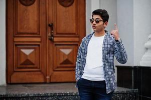 Stylish indian young man at sunglasses wear casual posed outdoor against door of building, show finger up, he has idea. photo