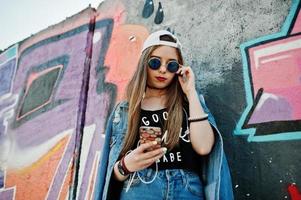 Stylish casual hipster girl in cap, sunglasses and jeans wear, listening music from headphones of mobile phone against large graffiti wall. photo