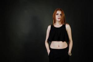 Portrait of a beautiful redheaded girl in black top and black skirt posing in the studio next to the grey wall. photo