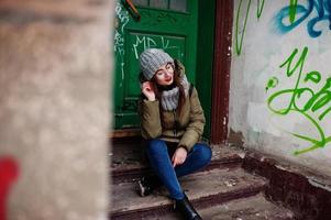 Portrait of brunette girl in gray scarf and hat, glasses sitting against urban entrance. photo