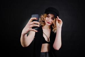 Studio portrait of blonde girl in black wear, bra and cap making selfie against dark background. photo