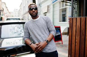 Stylish african american boy on gray sweater and glasses posed at street against black business car. Fashionable black guy. photo