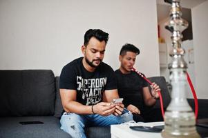 Two handsome and fashionable indian mans relaxing at room and smoking hookah. One of them looking at mobile phone. photo
